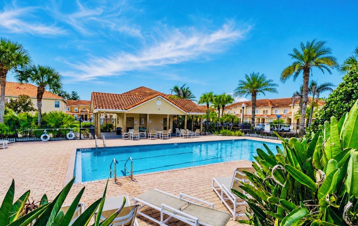 A swimming pool with lounge chairs, surrounded by palm trees and a building with a tiled roof.