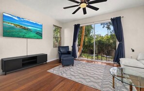 A living room with a flat-screen TV, a ceiling fan, a sofa, a glass coffee table, and a view of a bridge and greenery through the window.
