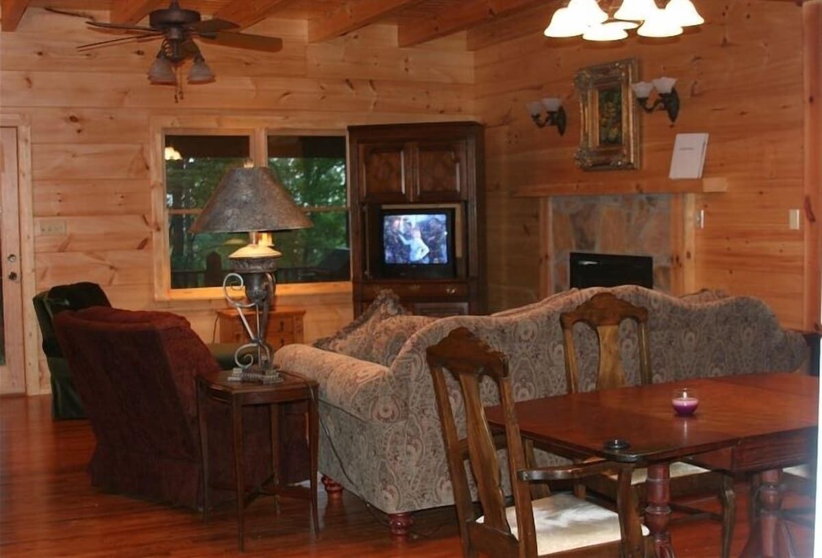 Acogedor interior de cabaña de madera con sofá, mesa de comedor, sillas, televisor y chimenea.