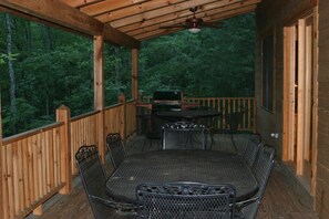 En overdækket terrasse med spisebord og stole, grill og udsigt til et skovområde.