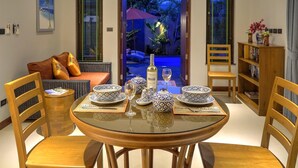A dining area with a round glass table set for four, surrounded by wooden chairs. A sofa with cushions is visible in the background, and a bookshelf with books and decorative items is to the right. - Private Villa with Swimming pool, jetted Plunge Pool and Free Transfers (Koh Samui)