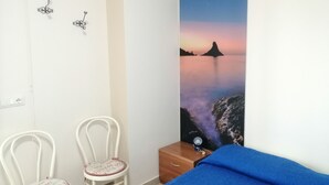 Un dormitorio con una cama, dos sillas, una mesita de noche y un cuadro colgado en la pared que muestra un atardecer sobre el agua.