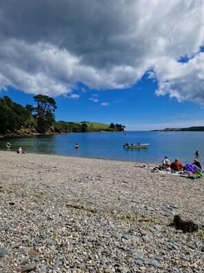 A pebbled beach with people sunbathing and swimming, boats on the water, and a lighthouse in the distance.