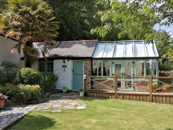 A quaint cottage with a green door, a stone pathway leading to a wooden deck, and a conservatory with glass walls.