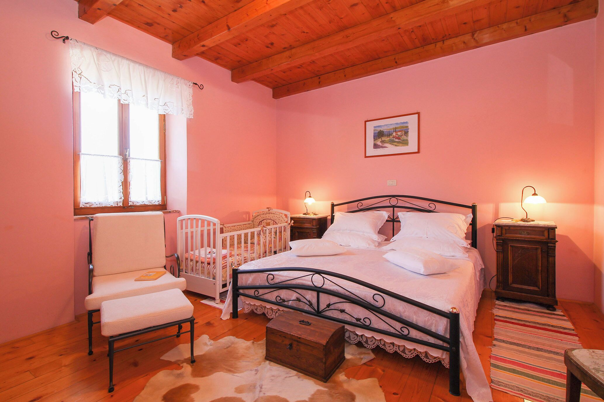 A bedroom with a wooden ceiling, a bed with white bedding, a chair, a crib, and a nightstand with a lamp.