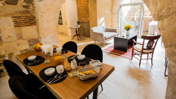 A dining area with a wooden table set for breakfast, stone walls, and a view of the outdoors. - Charming apartment in the heart of bergerac (Bergerac)