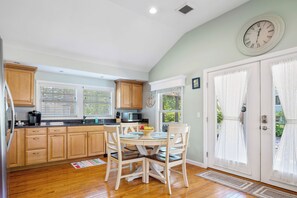A kitchen with wooden cabinets, a dining table with chairs, a microwave, and a clock on the wall. - ★The Islander | Steps From Water, Fire Pit, AC★ (Mashpee)