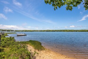 A lake with a dock, sandy shore, and a forested background. - ★The Islander | Steps From Water, Fire Pit, AC★ (Mashpee)