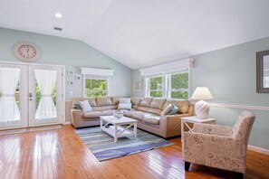 A living room with a sectional sofa, a coffee table, a patterned armchair, and a lamp. - ★The Islander | Steps From Water, Fire Pit, AC★ (Mashpee)