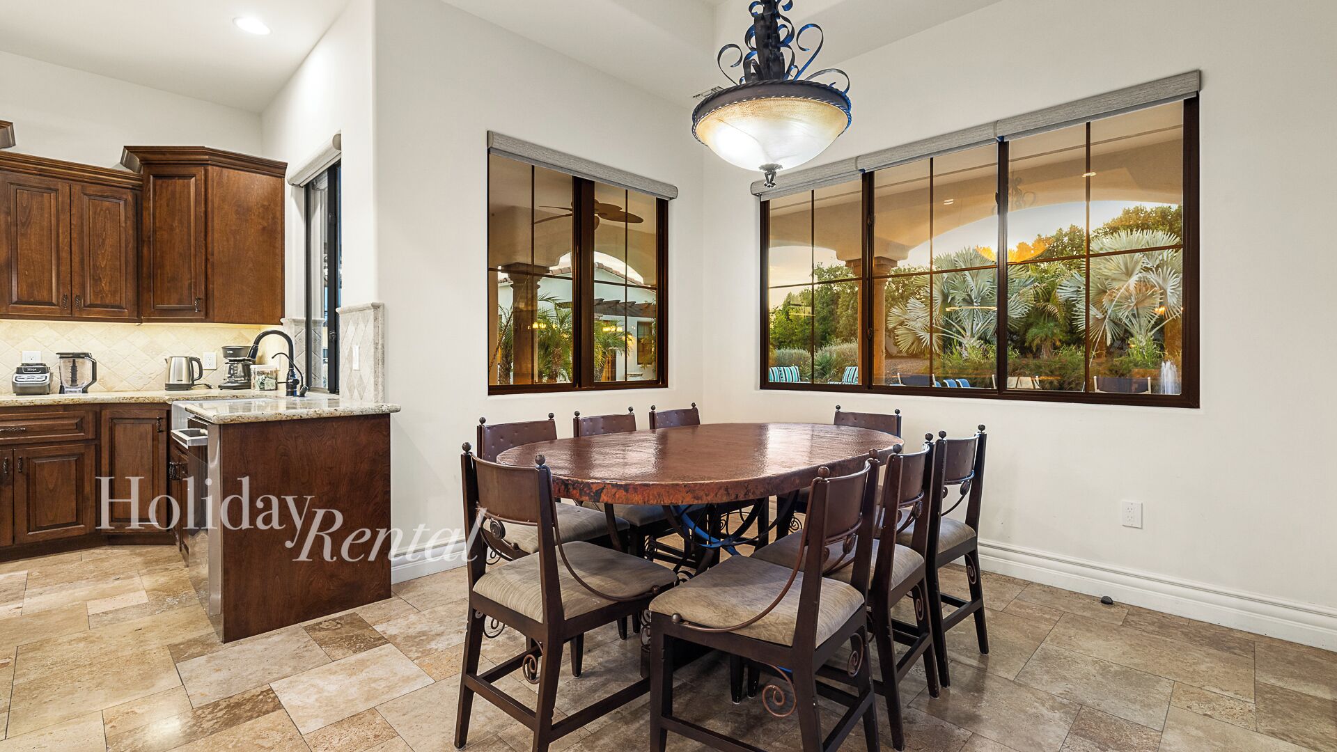 A spacious kitchen with dark wooden cabinets, a granite countertop, and a large island. There is a dining area with a round wooden table and six chairs. The room has large windows with a view of a garden.