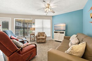 A living room with a ceiling fan, a television, and a view of the outdoors through large windows. - Popular beachside condo for 6 (Gulf Shores)
