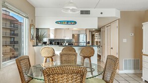 Een eethoek met een glazen tafel en rieten stoelen, een keuken met een magnetron en koelkast, en een uitzicht op een gebouw door een groot raam.