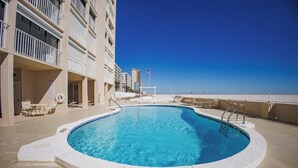 A swimming pool with a view of the ocean, surrounded by a building with balconies and a clear sky. - Beachfront condo with glass-enclosed balcony (Gulf Shores)