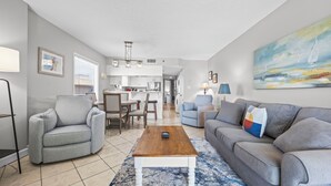 A living room with a grey sofa, a wooden coffee table, and a dining area with striped chairs.