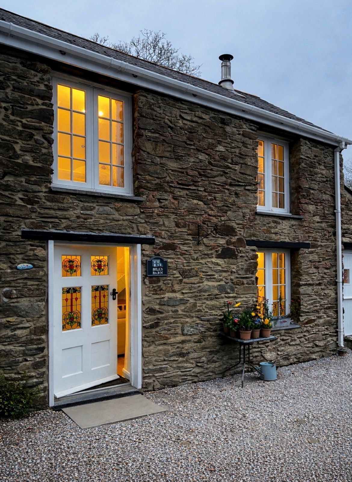 A stone building with a white door, windows with yellow lights, and a sign reading 'Hill's Barn'.