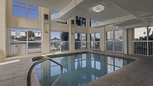 Une piscine intérieure avec des parois en verre transparent, un escalier métallique et une vue sur les palmiers et les bâtiments à l’extérieur.