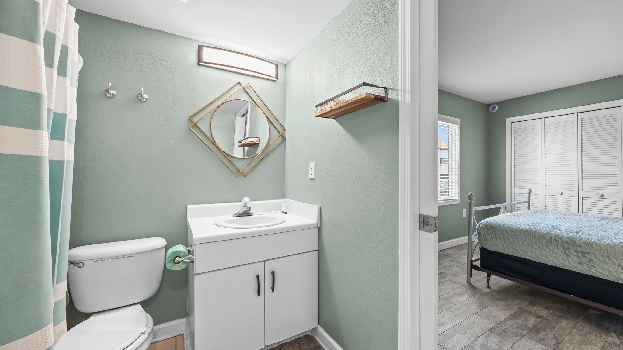 A bathroom with a toilet, sink, and mirror, and a bedroom with a bed and window.
