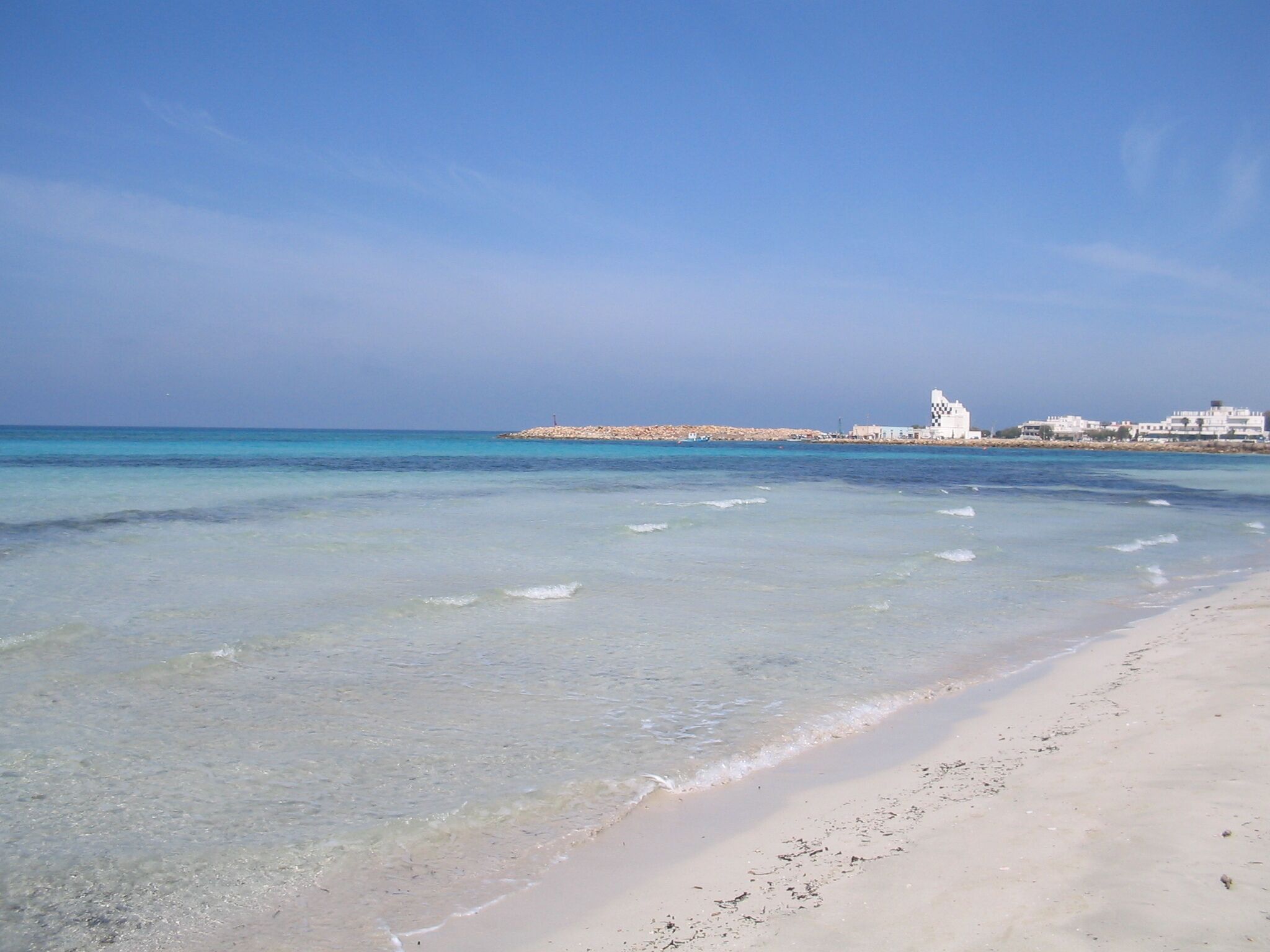 Ein Strand mit klarem, flachen Wasser, sandigem Ufer und Gebäuden im Hintergrund.