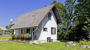 Ein weißes Haus mit Satteldach, einer kleinen Veranda und einem Garten mit Blumen und einer Bank.