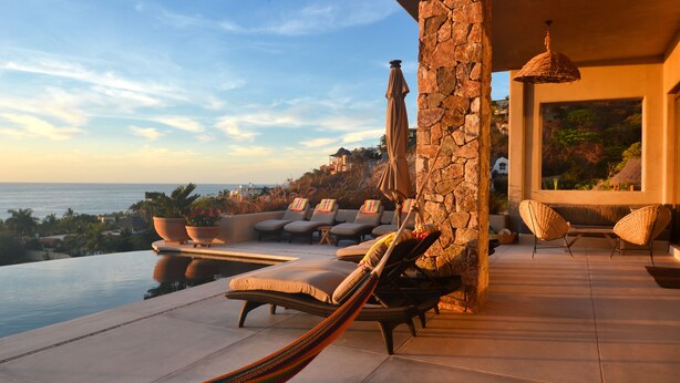 A luxurious outdoor lounge area with a stone wall, a hammock, and lounge chairs with cushions, overlooking a pool and the ocean. - 5 Star Luxe, Ocean Views, Walk to Beach & Village, Full Staff, 8-10 BR Rates (Sayulita)