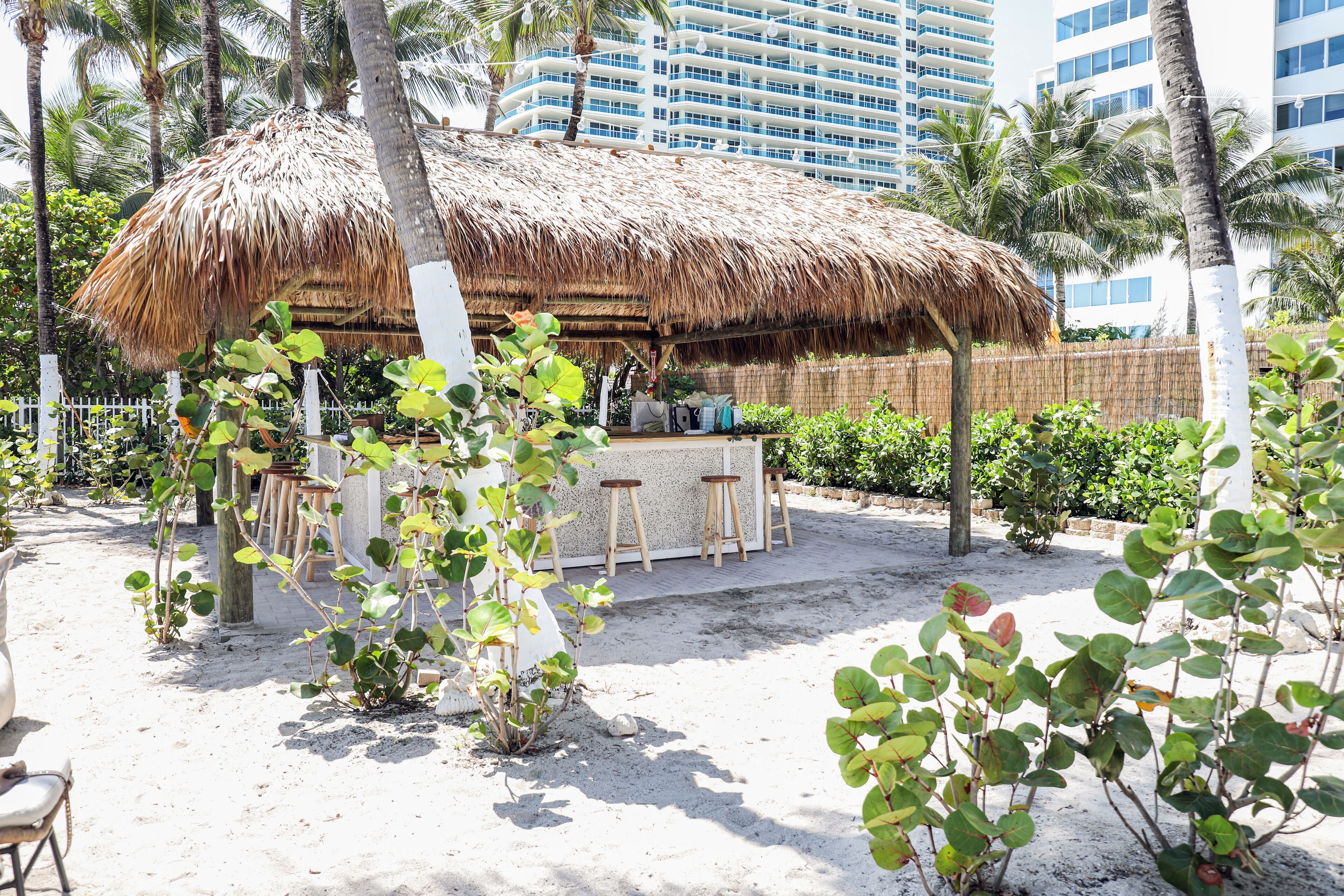A thatched-roof outdoor seating area with stools, surrounded by palm trees and plants, with a building in the background.
