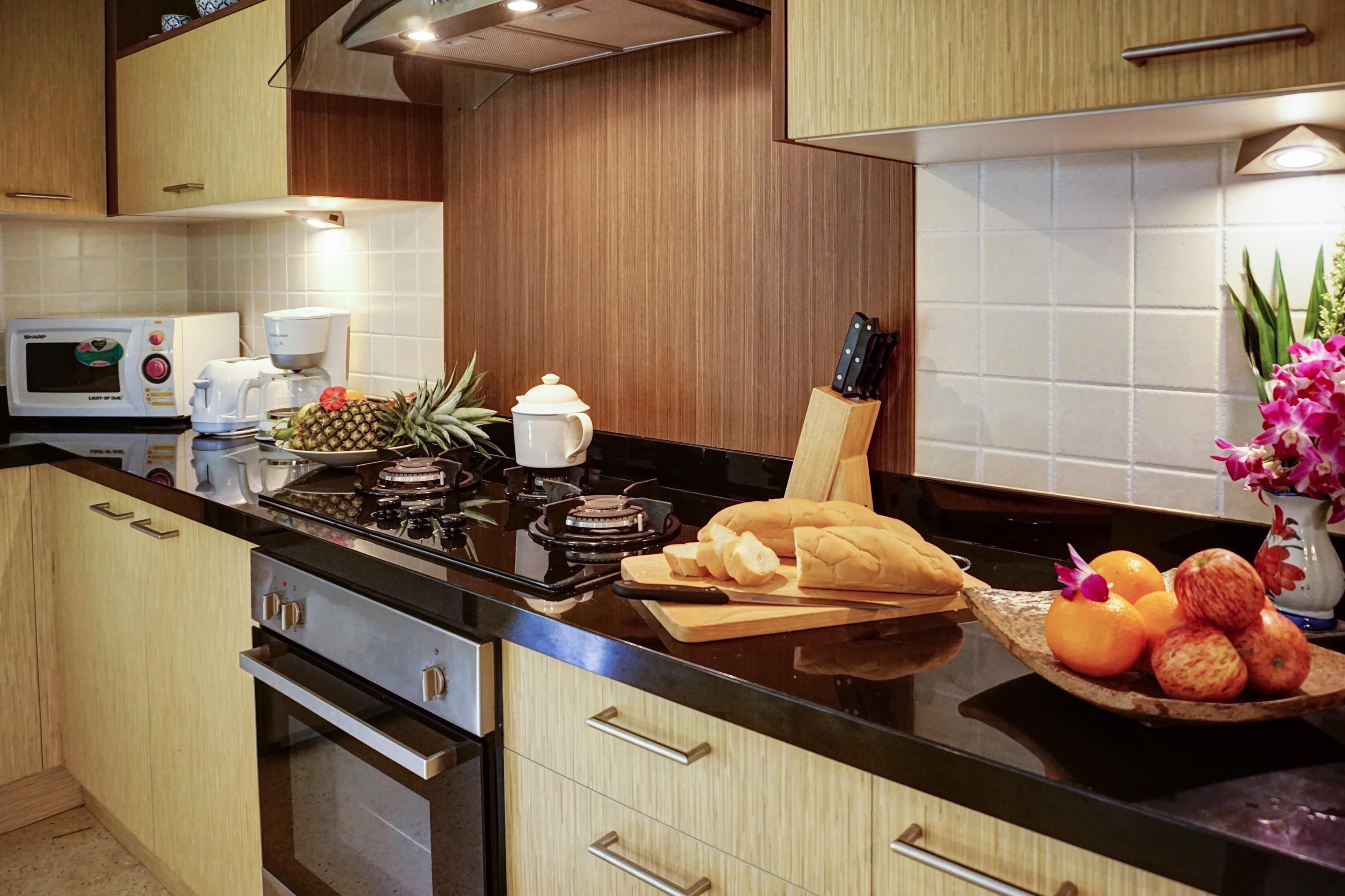 A modern kitchen with wooden cabinets, a black countertop, a microwave, a coffee maker, a cutting board, a knife, a bread loaf, and a bowl of fruit.