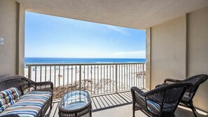Un balcón con mobiliario de mimbre y una mesa de vidrio, con vista a una playa con sombrillas y gente.