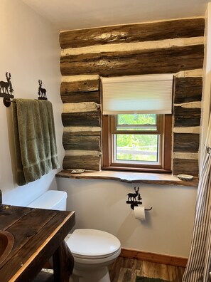 A rustic bathroom with a wooden log wall, a toilet, a sink with a wooden countertop, and a window with a view of greenery. - Cabin Cove on the river (Iowa Falls)