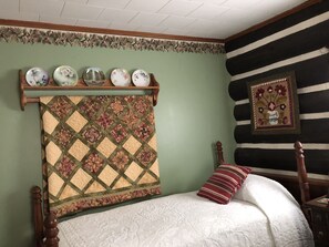 A bedroom with a bed, a quilt, a wooden headboard, a shelf with decorative plates, and a framed floral artwork. - Cabin Cove on the river (Iowa Falls)
