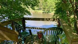 A wooden deck with two green chairs and a potted plant, overlooking a calm body of water and a grassy area.