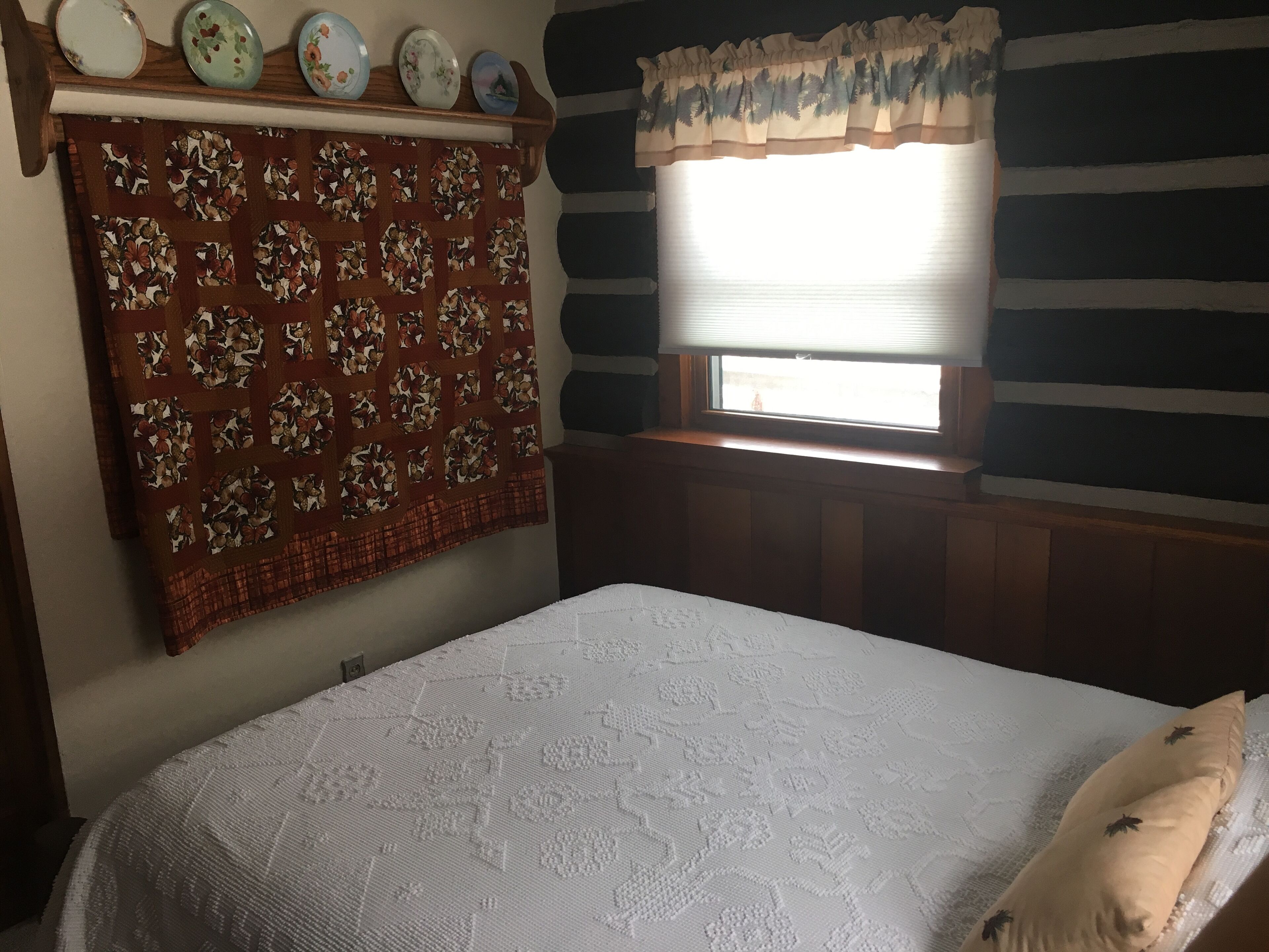 A bedroom with a bed, a quilt, a window with blinds, and a wall with a striped pattern.