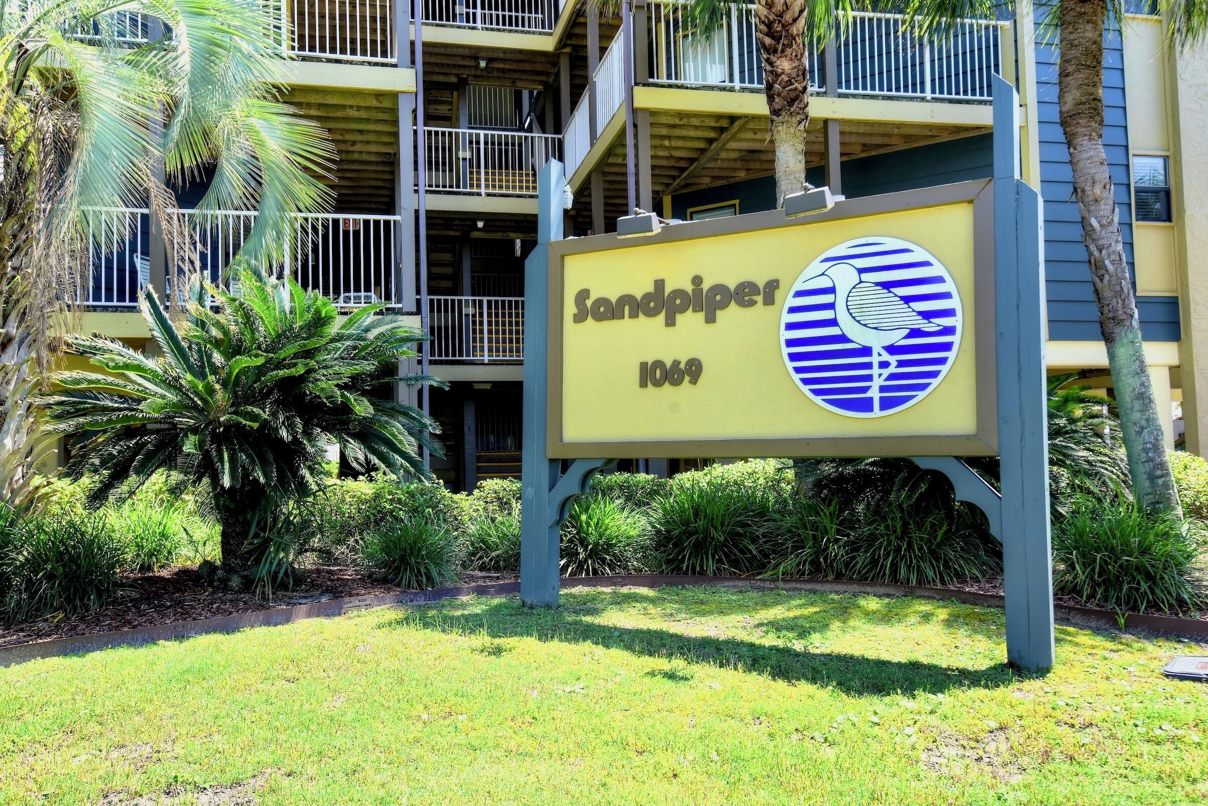 A sign for Sandpiper 1069 with a bird logo, surrounded by palm trees and a building with balconies.