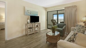 Un salón con televisión de pantalla plana, un sofá, una silla, una mesa de centro de vidrio y un amplio ventanal corredero que da a un balcón.
