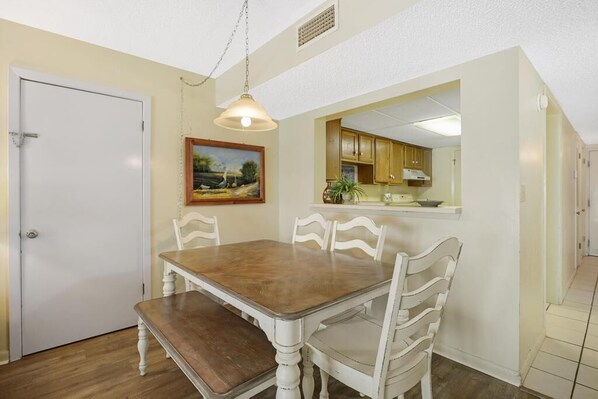 A dining room with a wooden table and white chairs, a painting on the wall, and a kitchen area visible through an archway.