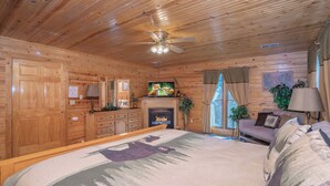 Una habitación tipo cabaña de madera con cama, chimenea, paredes de madera, ventilador de techo y televisor.