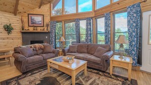 Un salón acogedor con dos sofás de cuero marrón, una mesa de centro de madera y una chimenea. Amplias ventanas con cortinas azules ofrecen vistas al exterior.
