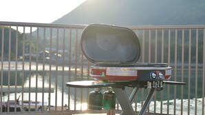 Una parrilla portátil con la tapa abierta, sobre una mesa con vista a un cuerpo de agua y montañas al fondo.
