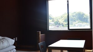 Una habitación con cama, una mesa y una silla. Se ve una ventana con vistas a un paisaje verde y edificios.