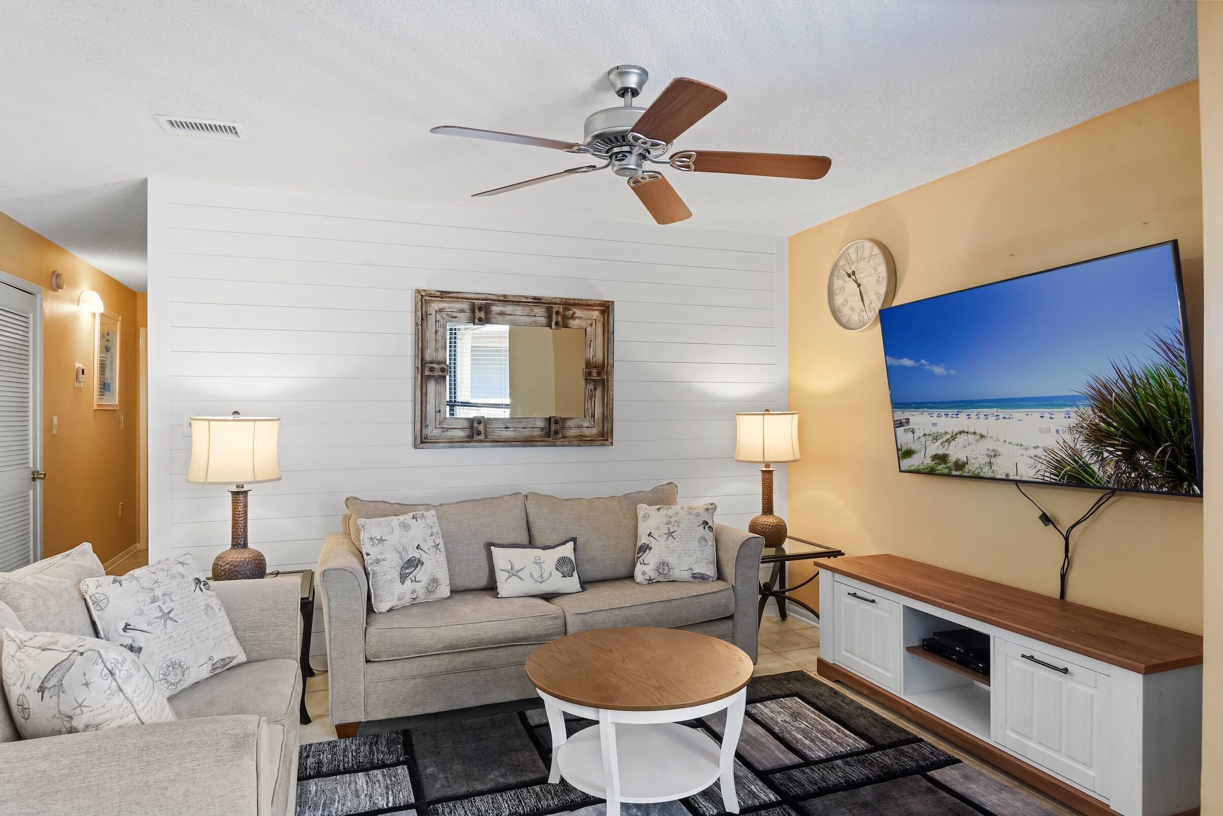 A living room with a beige sofa, a wooden coffee table, a TV mounted on the wall, and a ceiling fan.