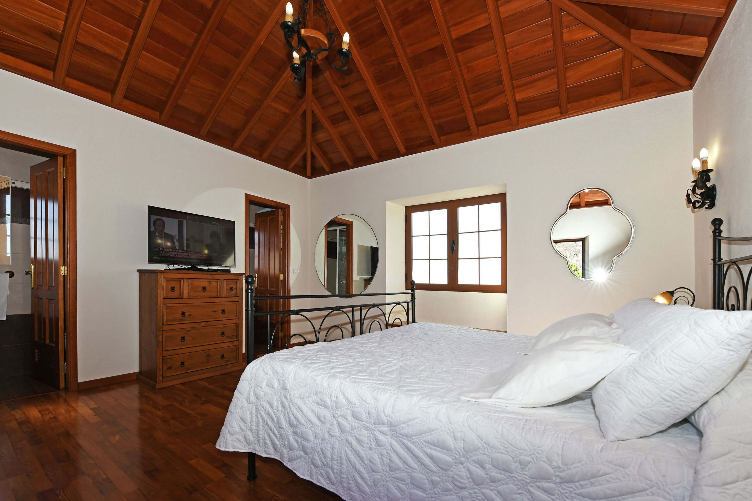 Une chambre avec un plafond en bois, un lit avec une literie blanche, une commode, un téléviseur et deux miroirs fixés au mur.