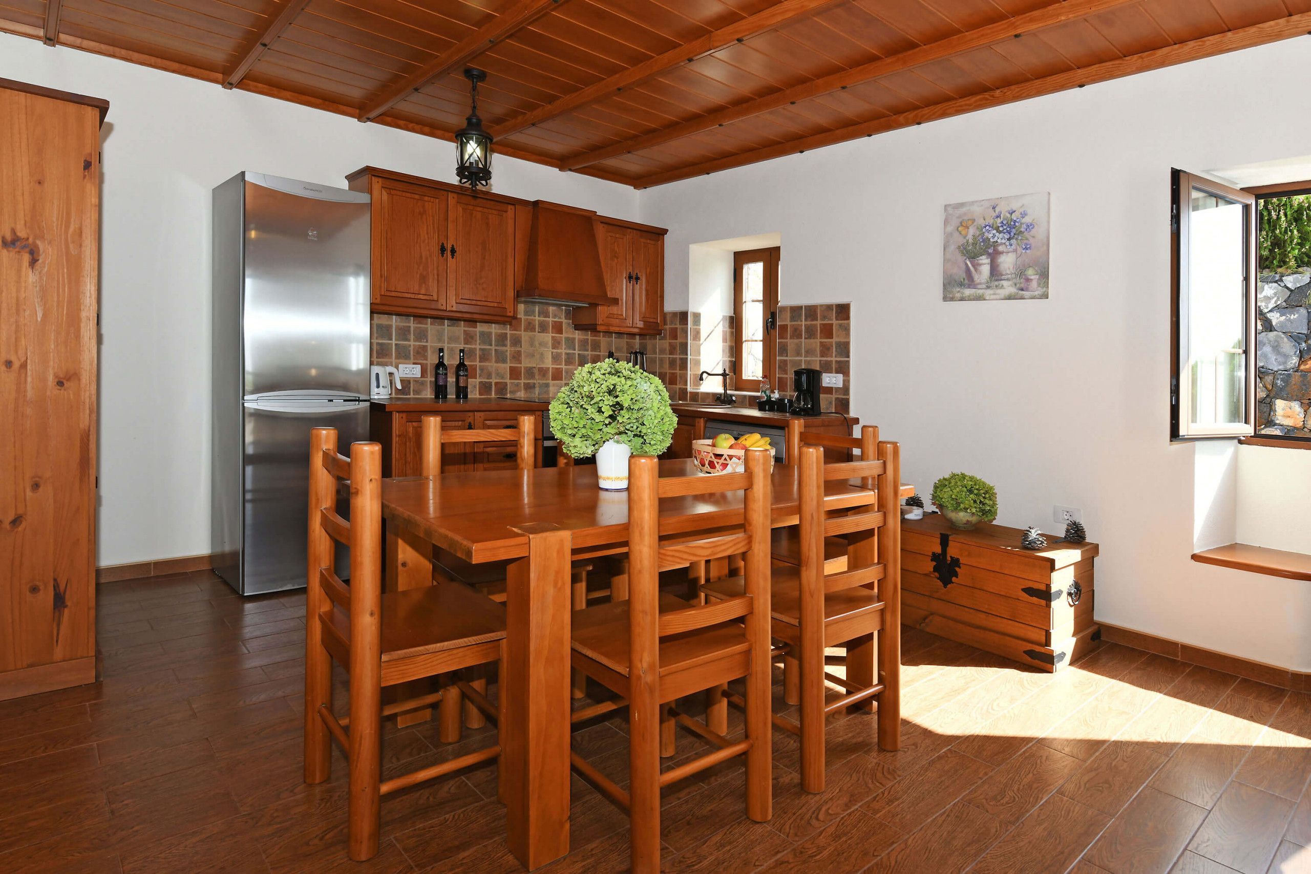 Une cuisine avec des meubles en bois, un réfrigérateur en acier inoxydable, une table à manger avec des chaises et une petite plante en pot.