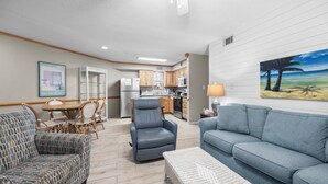 A compact living space with a dining area, a gray sofa, a wicker chair, a gray armchair, and a kitchen with wooden cabinets. - Sandpiper 1A ~ Family Fun ~ Comfortable ~ Corner Unit (Gulf Shores)