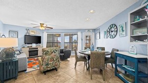 A spacious living room with a television, comfortable seating, a dining area, and a ceiling fan. - Sandpiper 7A ~ Enjoy the pool and beach views (Gulf Shores)