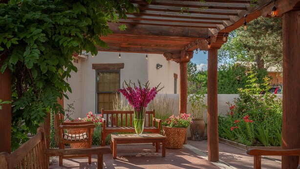 Una pérgola de madera con bancos y plantas en macetas, un edificio blanco al fondo y un cielo despejado.