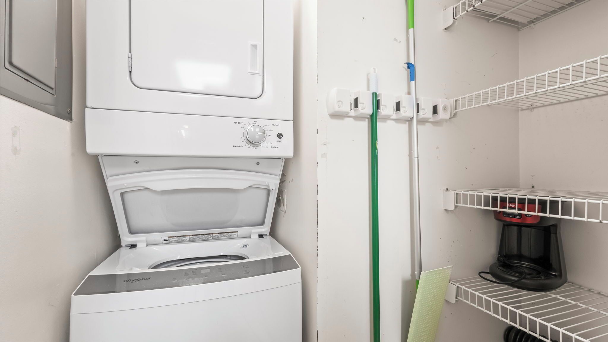 Un área de lavandería compacta con una lavadora Whirlpool de carga frontal, un tendedero y una cafetera sobre una repisa.