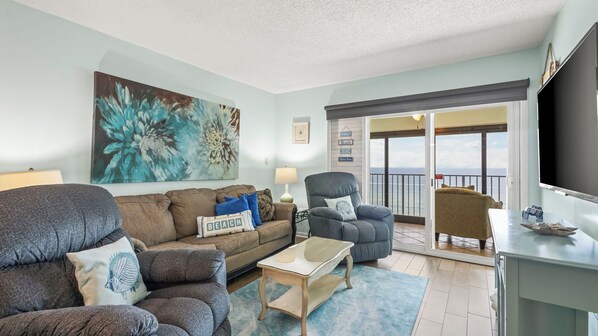 A living room with a sofa, armchair, coffee table, and a painting. There is a balcony with a view of the ocean.
