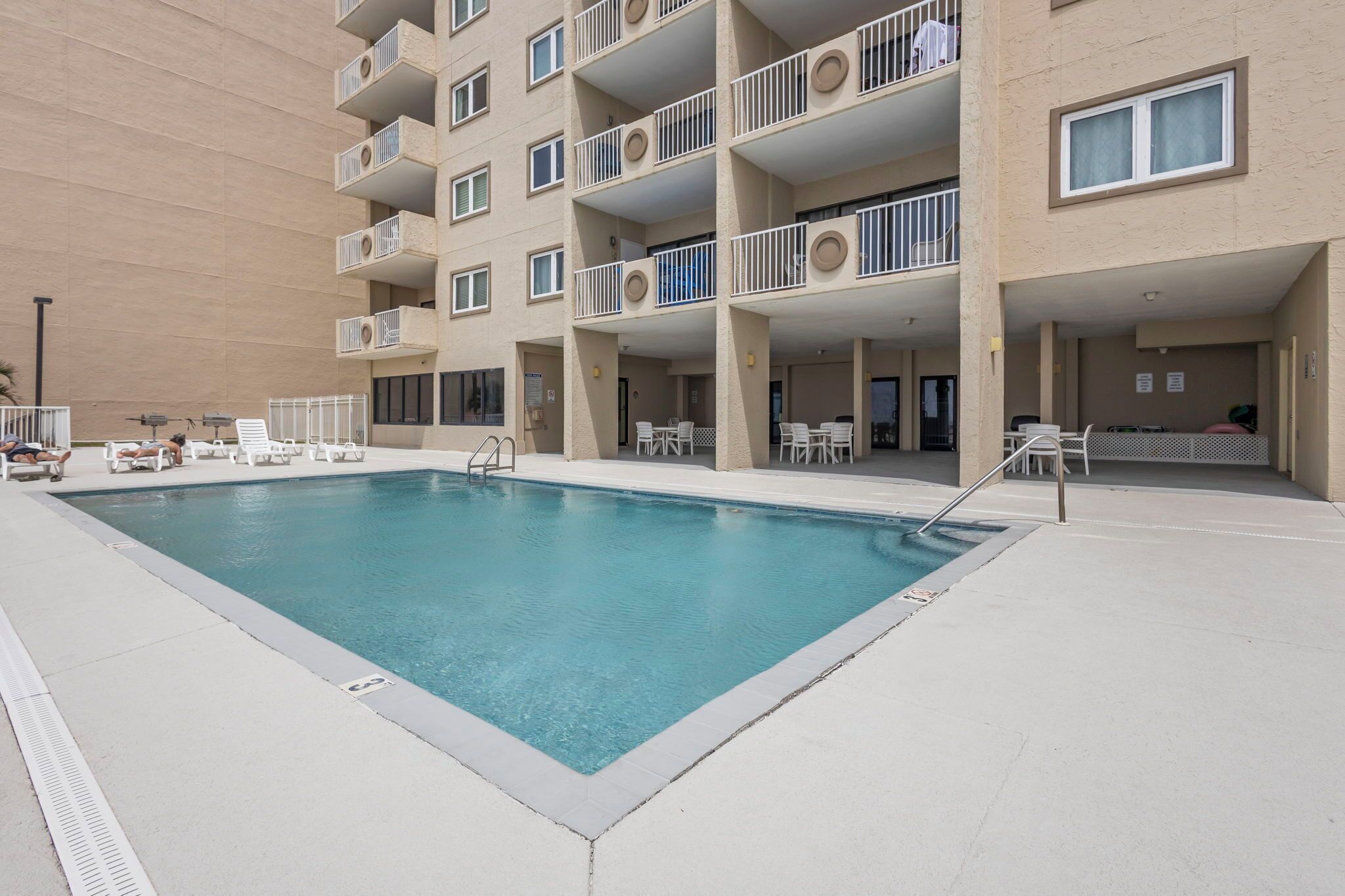 A swimming pool with lounge chairs and tables, surrounded by a multi-story building with balconies.