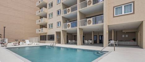 A swimming pool with lounge chairs and tables, surrounded by a multi-story building with balconies.