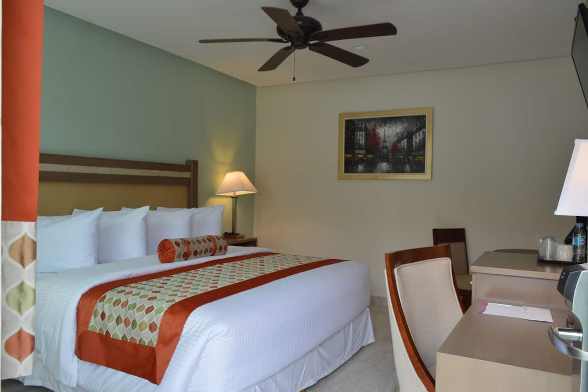 A hotel room with a bed, a desk, a chair, a ceiling fan, a lamp, and a painting on the wall.