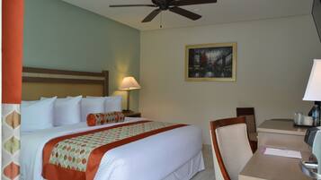 A hotel room with a bed, a desk, a chair, a ceiling fan, a lamp, and a painting on the wall.
